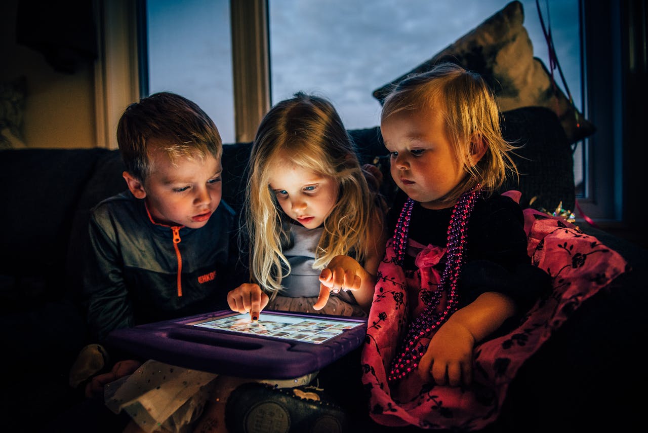 Contact Three young children engrossed in a tablet while sitting indoors, illuminated by the screen.