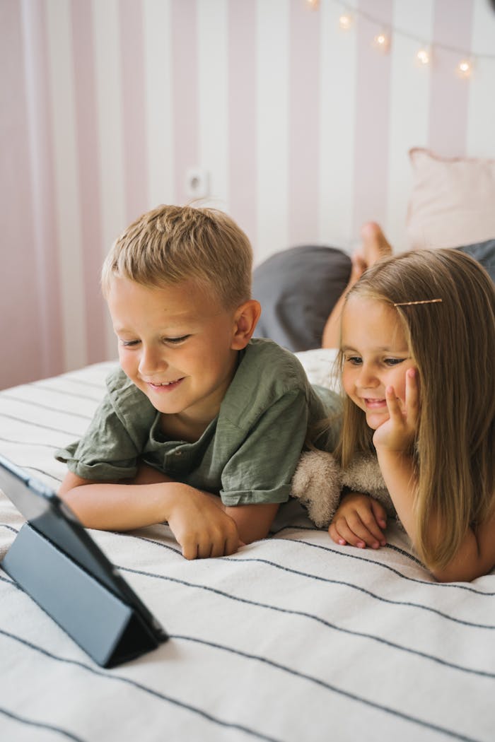 Two smiling children watching a tablet together on a bed, enjoying playful moments indoors.