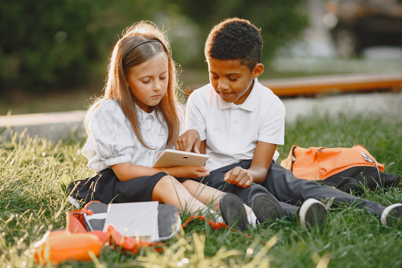 Two schoolchildren in uniforms using a tablet outdoors during golden hour.