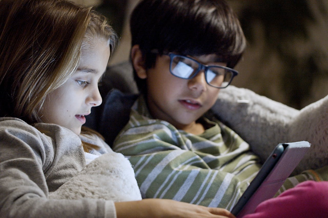 Startseite Two children comfortably using a tablet in bed at night in a cozy setting.