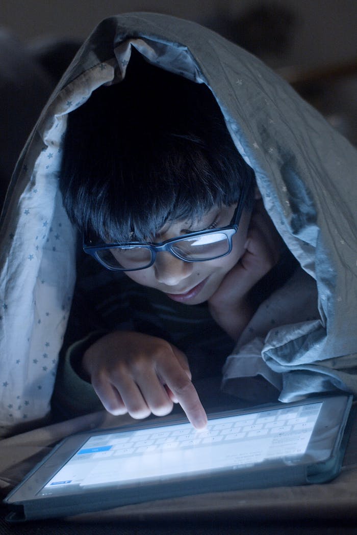 Kid using a digital tablet under a blanket at night, focused and attentive, in a cozy indoor setting.